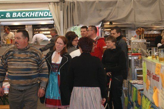 Eine Gruppe von Menschen steht in einem Imbiss, einige tragen Brillen und eine Frau trägt ein traditionelles Kleid. Ein Mann steht hinter dem Tresen und einige Leute stehen vor dem Tresen. Ein paar Flaschen stehen auf dem Tresen.
