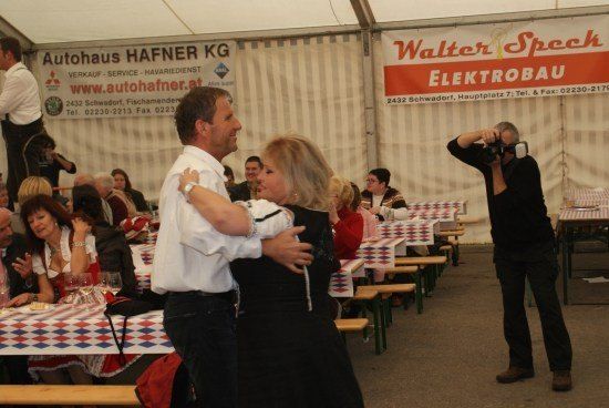 Ein Mann und eine Frau tanzen in einem Zelt mit Tischen und Stühlen, während ein Fotograf den Moment einfängt. Menschen sitzen an Tischen, und es gibt Banner, die Autohaus Hafner und Walter Spech Elektrobau bewerben.