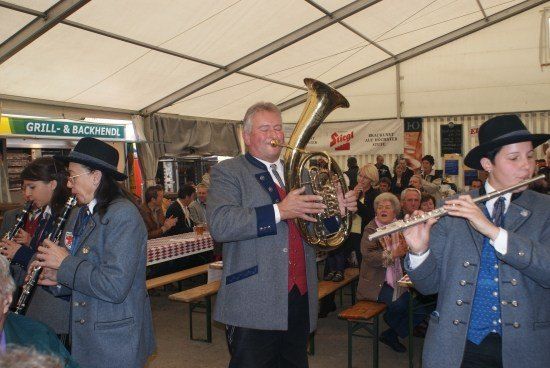 Eine Gruppe von Musikern, darunter ein Mann, der Tuba spielt, eine Frau, die Flöte spielt, und andere, die verschiedene Instrumente spielen, tritt in einem Zelt auf. Zuschauer sitzen an Tischen hinter ihnen.