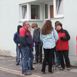 Eine Gruppe von Menschen in Winterkleidung steht vor einem Gebäude, möglicherweise warten sie auf etwas. Einige halten Skistöcke.
