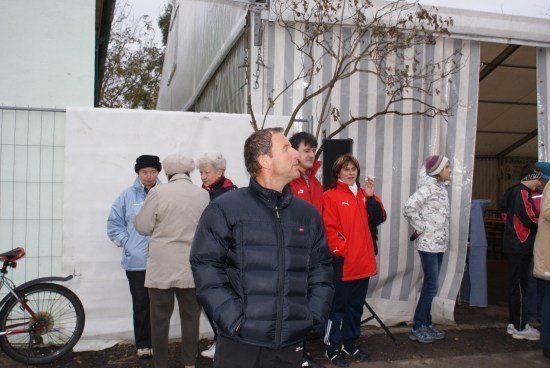 Eine Gruppe von Menschen in Winterkleidung steht vor einer weißen Wand mit einem Baumzweig und einem Fahrrad in der Nähe.