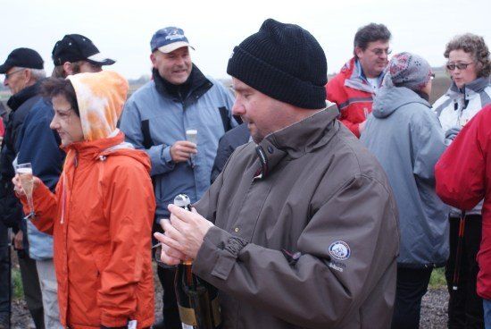 Eine Gruppe von Menschen in Winterkleidung steht draußen. Einige halten Gläser mit Champagner. Ein Mann in einem braunen Mantel öffnet eine Flasche.