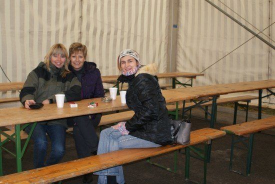 Drei Frauen sitzen an einem Holztisch in einem Zelt, lächeln und posieren für ein Foto. Sie haben Tassen und ein Handy auf dem Tisch.