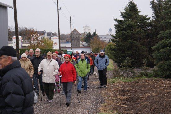 Eine Gruppe von Menschen geht auf einem Kiesweg in einem ländlichen Gebiet, einige mit Stöcken. Bäume und ein Gebäude sind im Hintergrund.