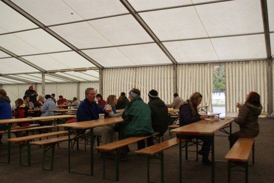 Eine Gruppe von Menschen sitzt unter einem großen Zelt an Tischen, einige essen und andere unterhalten sich. Das Zelt ist weiß mit Metallstützen und es gibt Fenster an der Seite.