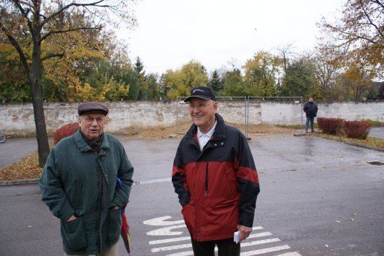 Zwei ältere Männer stehen auf einem Parkplatz mit einer weißen Mauer und Bäumen im Hintergrund. Ein Mann trägt eine grüne Jacke, der andere eine rote und schwarze Jacke.