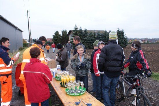 Eine Gruppe von Menschen, einige in orangefarbenen Sicherheitsanzügen, versammelt sich um einen Tisch mit Getränken und einem Fahrrad in einer ländlichen Umgebung.
