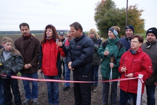 Eine Gruppe von Menschen steht um ein Band, möglicherweise für eine Bandzeremonie. Sie sind in Winterkleidung gekleidet und einige halten Gläser. Eine Frau in einer roten Jacke hält ein Mikrofon.