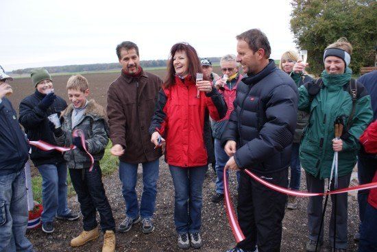 Eine Gruppe von Menschen, darunter Erwachsene und Kinder, stehen auf einem Feld, halten eine Schleife und lächeln. Sie feiern wahrscheinlich eine Schleifenschneidezeremonie.
