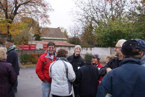 Eine Gruppe von Menschen, darunter ein Mann in einer roten Jacke, stehen im Kreis in einem Außenbereich. Sie sind von Bäumen und einem Gebäude umgeben.