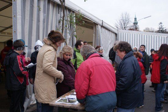 Eine Gruppe von Menschen, einige in Wintermänteln, stehen um einen Tisch mit Papieren und einer Karte herum, möglicherweise warten sie in einer Schlange vor einem Zelt.