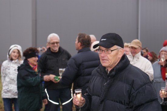 Ein älterer Mann in einer schwarzen Daunenjacke und Kappe spricht zu einer Menschenmenge, einige halten Champagnerflöten, im Freien.