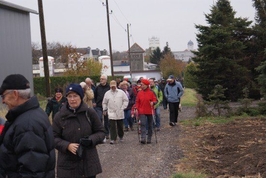 Eine Gruppe von Menschen geht auf einem Kiesweg, einige tragen Mützen und tragen Stöcke, mit Bäumen und Gebäuden im Hintergrund.
