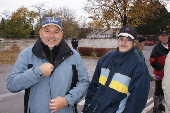 Zwei Männer in Jacken stehen auf einer Straße, einer trägt einen blauen Hut und der andere einen schwarzen Hut mit weißer Kante. Sie lächeln und scheinen für ein Foto zu posieren. Dahinter befindet sich eine Mauer mit einem Gebäude und Bäumen.