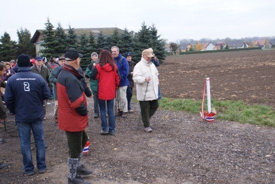 Eine Gruppe von Menschen ist auf einem Kiesweg versammelt, einige tragen Jacken und Mützen. Im Hintergrund ist ein Feld mit Bäumen und einem Haus zu sehen.