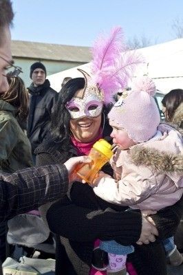 Eine Frau mit Maske hält ein Baby in den Armen, lächelt und hält eine gelbe Flasche. Eine Person in einem karierten Hemd hält die Hand des Babys.