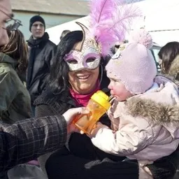 Eine Frau mit Maske hält ein Baby in den Armen, lächelt und hält eine gelbe Flasche. Eine Person in einem karierten Hemd hält die Hand des Babys.