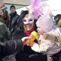 Eine Frau mit Maske hält ein Baby in den Armen, lächelt und hält eine gelbe Flasche. Eine Person in einem karierten Hemd hält die Hand des Babys.