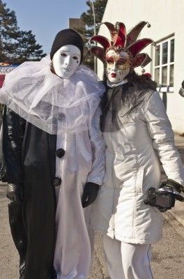 Zwei Personen in aufwendigen Kostümen, eine in Schwarz und Weiß mit weißer Maske, die andere in Weiß mit roter und schwarzer Maske, stehen zusammen auf einer Straße.