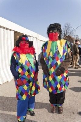 Zwei Personen in farbenfrohen Harlekin-Kostümen stehen nebeneinander im Freien. Beide tragen rote Masken und Handschuhe, einer eine Mütze. Ein weißes Zelt und Menschen im Hintergrund deuten auf eine festliche Veranstaltung hin.