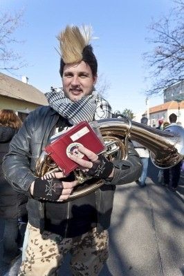 Ein Mann mit einem Irokesenschnitt und einem Schal hält ein rotes Buch und eine Tuba in der Straße.