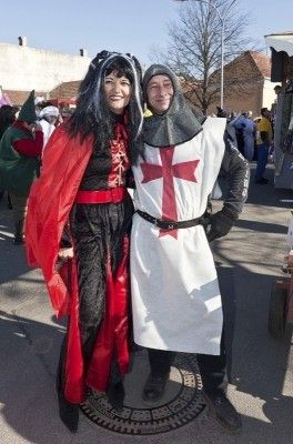 Eine Frau in einem roten Umhang und ein Mann in einem weißen Ritterkostüm mit einem roten Kreuz stehen zusammen auf einer Straße während eines Festivals.