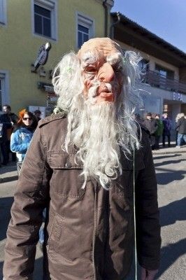 Eine Person mit brauner Jacke und weißer Bartmaske steht auf einer Straße mit Gebäuden und Menschen im Hintergrund.