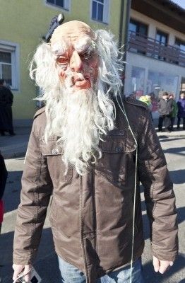 Eine Person in einer braunen Jacke und einer weißen Perücke, mit einer Maske, die einem alten Mann ähnelt, geht auf der Straße.