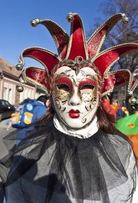 Eine Person, die eine rote und goldene Narrenmaske mit weißem Gesicht trägt, steht auf einer Straße mit Gebäuden und Autos im Hintergrund.