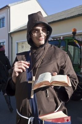 Ein Mann, der als Mönch verkleidet ist, hält ein offenes Buch und ein Glas Wein in den Händen. Er trägt einen braunen Umhang und eine Kapuze.
