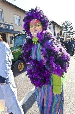 Eine Person in einem farbenfrohen Kostüm mit lila Federn und Gesichtsbemalung steht während eines Umzugs auf der Straße.