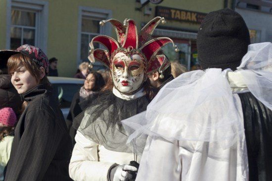Eine Person in einem weißen Kostüm mit einer rot-goldenen Maske steht unter anderen auf einer Straße, möglicherweise bei einem Fest.