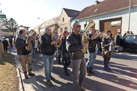 Eine Gruppe Musiker mit bunten Haaren und einzigartigen Outfits spielt Blechblasinstrumente, während sie auf einer Straße marschieren.