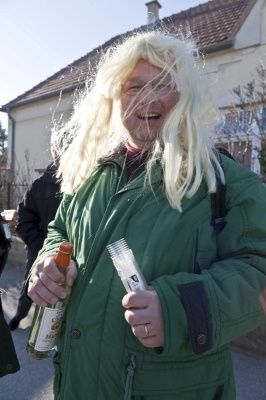 Ein Mann mit langen blonden Haaren in einer grünen Jacke hält eine Flasche und einen Plastikbecher vor einem Haus.
