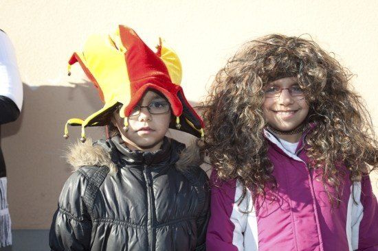 Zwei Kinder in Kostümen, eines in einem roten und gelben Narrenhut, das andere in einer pinken Jacke mit lockigem Haar und Brille.