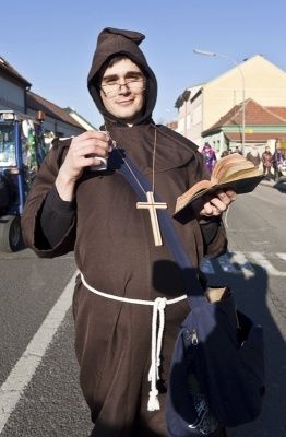 Ein Mann, der als Mönch verkleidet ist, hält ein Buch und ein Glas Wasser in der Hand und steht auf einer Straße mit Gebäuden und einem Fahrzeug im Hintergrund.