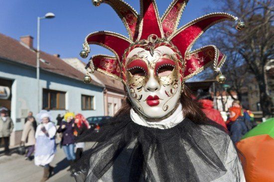 Eine Person in einer roten und goldenen Narrenmaske und einem schwarzen Spitzenkleid geht bei einer Parade. Dahinter sind Menschen in verschiedenen Kostümen und Gebäude zu sehen.