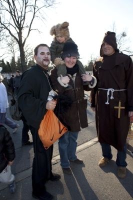 Drei Menschen in religiöser Kleidung stehen auf einer Straße, einer hält ein Kind und ein anderer einen Beutel. Sie sind von einer Menschenmenge umgeben, die auf dem Bürgersteig geht.