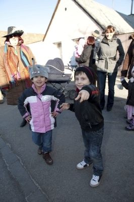 Zwei junge Jungen stehen auf einer Straße, einer hält eine Kamera und zeigt sie auf den anderen. Hinter ihnen steht eine Gruppe von Menschen, darunter eine Frau in einem Hut, in der Nähe eines Gebäudes.