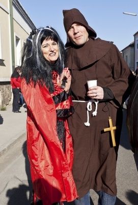 Ein Mann in Mönchskutte und eine Frau in rotem Kostüm stehen zusammen auf der Straße, der Mann hält einen Becher und eine Kreuzkette.
