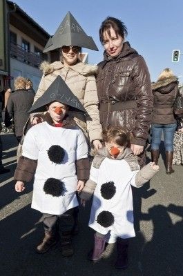 Drei Menschen in Winterkleidung, zwei davon in Schneemannkostümen, stehen auf einer Straße mit Gebäuden und Menschen im Hintergrund.