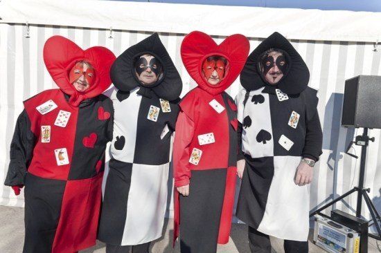 Vier Personen in Narrenkostümen mit herzförmigen Kapuzen und Gesichtsmasken, verziert mit Spielkarten-Designs, stehen zusammen vor einem weißen Zelt.