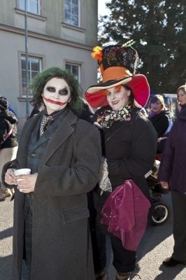 Zwei Personen in Kostümen, eine als Joker und die andere als Hutmacher, stehen im Freien mit einer Menschenmenge im Hintergrund.