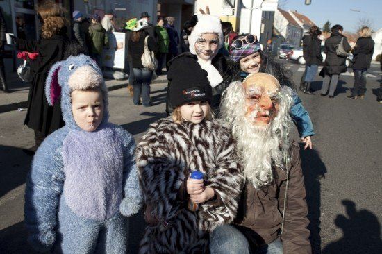 Eine Gruppe von Menschen in Kostümen, darunter ein Kind in einem blauen Eselkostüm, ein Kind in einem schwarzen und weißen Tigerkostüm und ein Erwachsener in einem weißen Bart- und Langhaarkostüm, posiert für ein Foto auf der Straße.