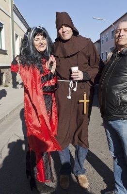 Drei Personen in Kostümen stehen auf einer Straße, eine von ihnen hält einen Becher und ein Kreuzanhänger.