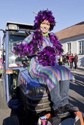 Eine Person in einem bunten Kostüm und einem lila Boa sitzt auf einem blauen Traktor und lächelt in die Kamera. Im Hintergrund stehen mehrere Personen und beobachten.