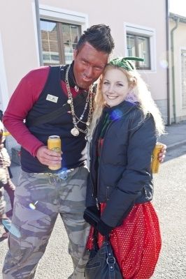Ein Mann und eine Frau in festlicher Kleidung stehen nebeneinander, halten Getränke in den Händen und posieren für ein Foto vor einem Gebäude.