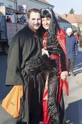 Ein Mann und eine Frau in Kostümen stehen auf der Straße, mit einem Lastwagen und Menschen im Hintergrund.