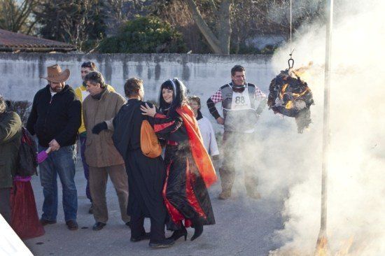 Eine Gruppe von Menschen versammelt sich um eine brennende Puppe. Eine Frau in einem roten und schwarzen Kostüm steht mit einem Mann in einem schwarzen Anzug.