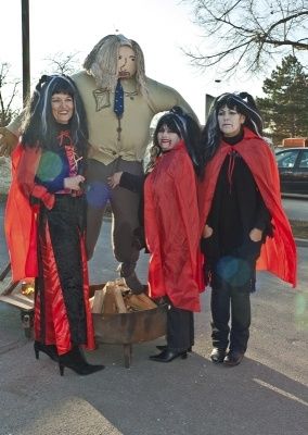 Vier Personen in roten Umhängen und schwarzen Outfits stehen vor einer großen Gandalf-Statue, möglicherweise für eine Halloween-Veranstaltung.